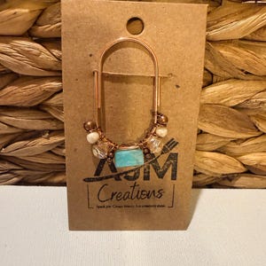 May include: A copper-colored earring with a rectangular turquoise stone and white beads. The earring is attached to a brown card with the text "AJM Creations" and the tagline "Spark joy. Create beauty. Let creativity shine."