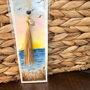 May include: A watercolor bookmark featuring a beach scene with a sunset, blue water, and flying birds. The bookmark has a gold tassel, a blue bead, and a small metal charm. The bookmark is encased in clear plastic.