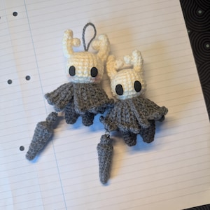 May include: Two handmade crochet plush figures featuring off-white heads with horn-like protrusions, large black circular eyes, and grey cloaks. Each figure is attached to a small, grey, cone-shaped accessory by a short string, and one has a loop.