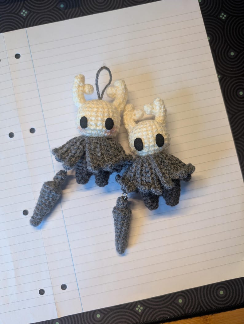 May include: Two handmade crochet plush figures featuring off-white heads with horn-like protrusions, large black circular eyes, and grey cloaks. Each figure is attached to a small, grey, cone-shaped accessory by a short string, and one has a loop.