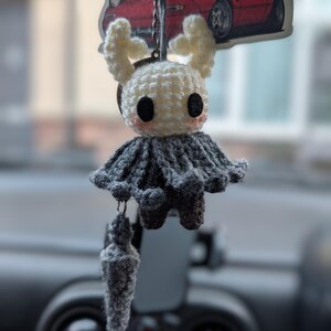 May include: A crochet keyring featuring a white, horned character with large black eyes and pink cheeks, wearing a dark grey cloak. A small, matching grey crochet nail-shaped accessory hangs from the bottom, all attached to a metal ring.