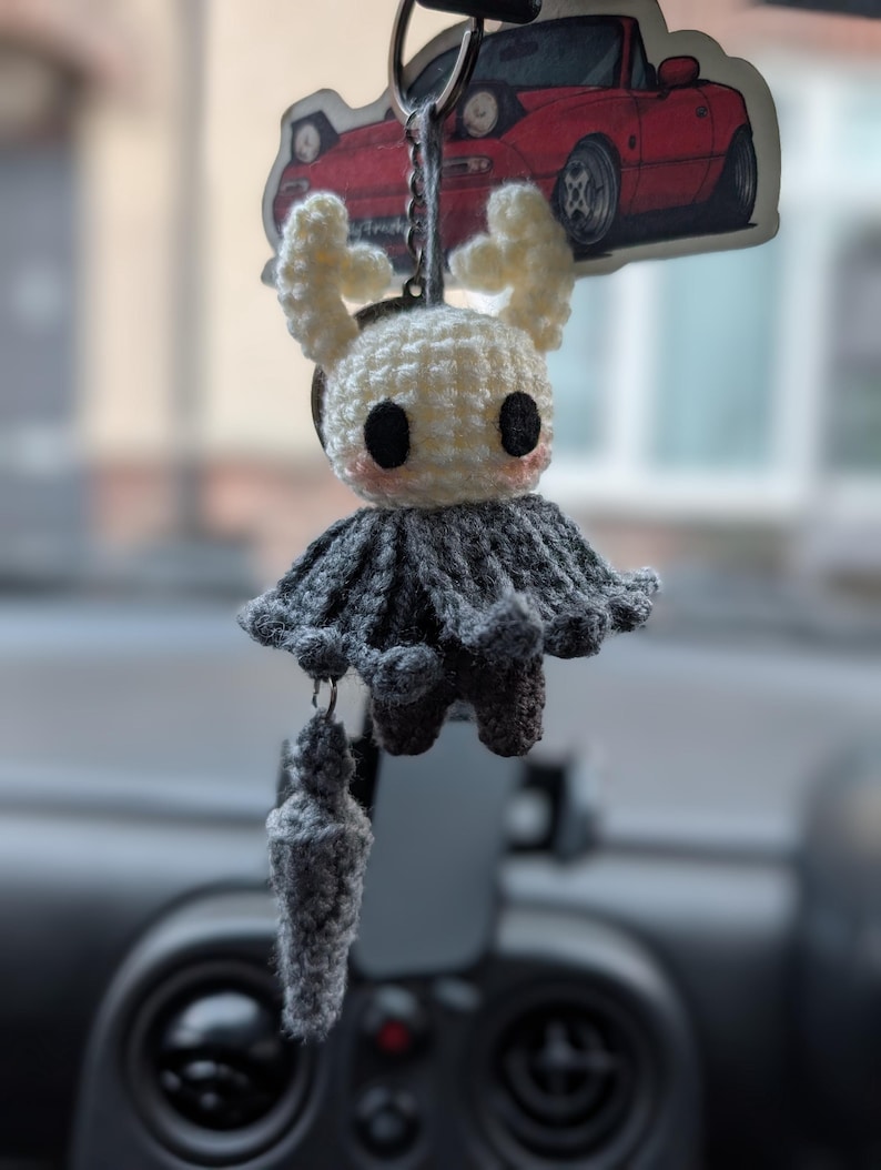 May include: A crochet keyring featuring a white, horned character with large black eyes and pink cheeks, wearing a dark grey cloak. A small, matching grey crochet nail-shaped accessory hangs from the bottom, all attached to a metal ring.