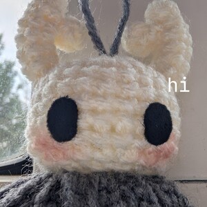 May include: A handmade crochet plush toy featuring a white head with two large black circular eyes, pink blush accents, and two curved white horns. A grey loop is attached to the top. The word hi is printed in white on the right side of the head.