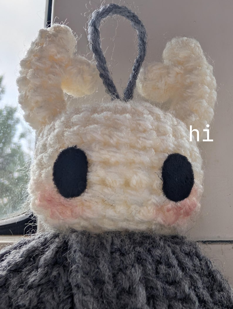 May include: A handmade crochet plush toy featuring a white head with two large black circular eyes, pink blush accents, and two curved white horns. A grey loop is attached to the top. The word hi is printed in white on the right side of the head.