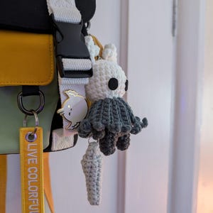 May include: A crochet plush keychain featuring a white head with black eyes and a dark grey cloak, attached to a yellow fabric tag with the text LIVE COLORFULLY and a small white bird-shaped enamel pin on a white strap of a black and yellow bag.