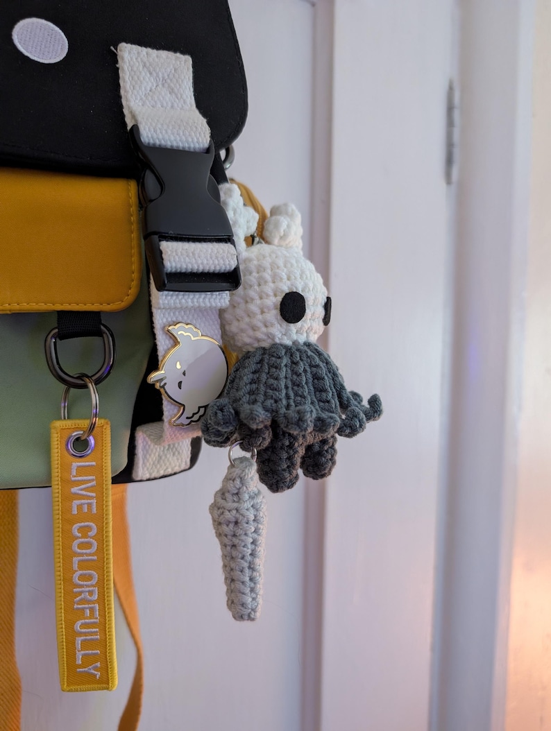 May include: A crochet plush keychain featuring a white head with black eyes and a dark grey cloak, attached to a yellow fabric tag with the text LIVE COLORFULLY and a small white bird-shaped enamel pin on a white strap of a black and yellow bag.