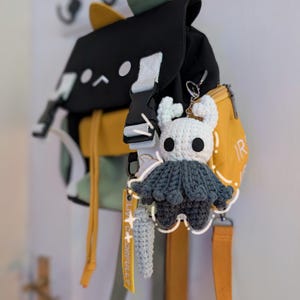 May include: A black, yellow, and light green backpack featuring a cat-ear design on the top flap. Attached to the bag is a white and dark grey crocheted character keychain with horns and a cape, alongside a small light blue crocheted accessory.