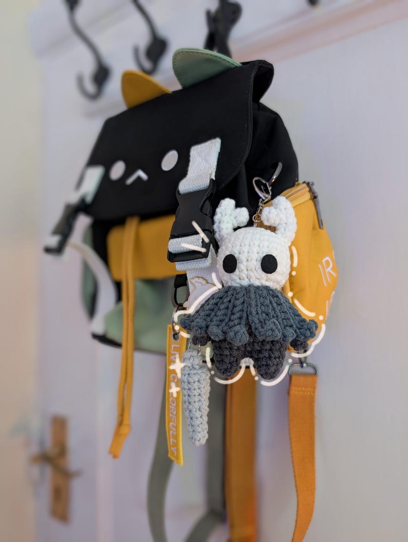 May include: A black, yellow, and light green backpack featuring a cat-ear design on the top flap. Attached to the bag is a white and dark grey crocheted character keychain with horns and a cape, alongside a small light blue crocheted accessory.
