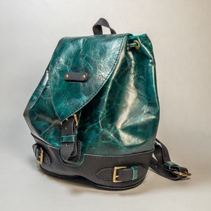 Puede incluir: Una mochila de cuero verde azulado con correas y hebillas negras. La mochila tiene un cierre de solapa y un aspecto de cuero desgastado.