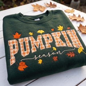 Könnte beinhalten: Ein dunkelgrüner Rundhals-Pullover mit dem Wort PUMPKIN in großen, orange-weiß karierten Blockbuchstaben. Darunter ist das Wort season in weißer Schreibschrift gestickt, umgeben von verschiedenen gestickten Herbstblättern.