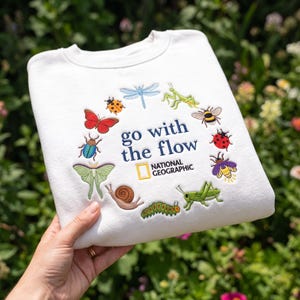 May include: A white crewneck sweatshirt featuring an embroidered circular arrangement of various insects including a dragonfly, mantis, bee, ladybug, firefly, grasshopper, caterpillar, snail, moth, beetle, and butterfly, with the text "go with the flow" and the National Geographic logo.