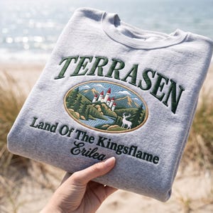 Puede incluir: Una sudadera gris claro de cuello redondo con bordado en verde oscuro que dice TERRASEN, Land Of The Kingsflame, Erilea. Un parche ovalado muestra un castillo blanco con techos rojos, montañas, pinos, un río y un ciervo blanco.