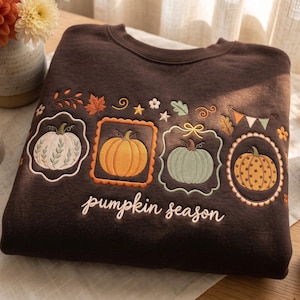 May include: A dark brown crewneck sweatshirt featuring embroidered designs of four distinct pumpkins in white, orange, sage green, and yellow. The embroidery includes autumn leaves, stars, and a bunting banner, with the text pumpkin season below.