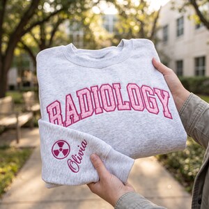Puede incluir: Una sudadera de cuello redondo en color gris jaspeado claro con la palabra RADIOLOGY bordada en una tipografía rosa de lunares en el pecho. La manga presenta un símbolo de radiación rosa bordado y el nombre Olivia en letra cursiva.