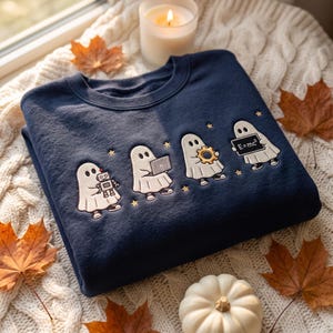 May include: A navy blue crewneck sweatshirt featuring an embroidered design of four white ghosts. The ghosts are depicted holding a small robot, a laptop, a golden gear, and a chalkboard with the text E=mc2 written in white, accented by stars.