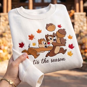 May include: An off-white crewneck sweatshirt featuring an embroidered design of two chipmunks holding acorns, surrounded by scattered red, orange, and yellow maple leaves. The text 'tis the season' is embroidered in dark brown cursive script below.