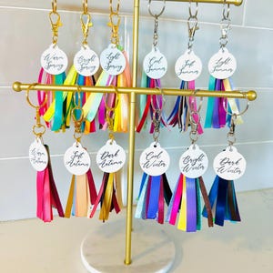 Pode incluir: Um expositor de metal dourado com chaveiros. Cada chaveiro tem uma etiqueta branca com nomes de paletas de cores sazonais como "Warm Spring" e uma borla colorida. As borlas são feitas de fitas de várias cores.