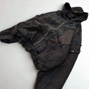 May include: Dark gray distressed hoodie and matching pants set. The hoodie features a zip-up front, a hood, and a patchwork design with frayed edges. The pants have a drawstring waist and a similar patchwork design. The set is made of a soft, comfortable material.