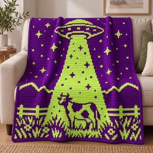 May include: A purple and lime green crochet blanket featuring a large UFO hovering above a cow in a field. The design includes a bright green tractor beam, stars, a wooden fence, and grass, all rendered in a pixelated, textured yarn pattern.