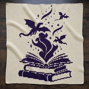 May include: A square crochet blanket in cream with a dark purple graphic design. The pattern features an open book at the bottom with a plume of smoke or magic rising from the pages, surrounded by several small flying dragon silhouettes and stars.