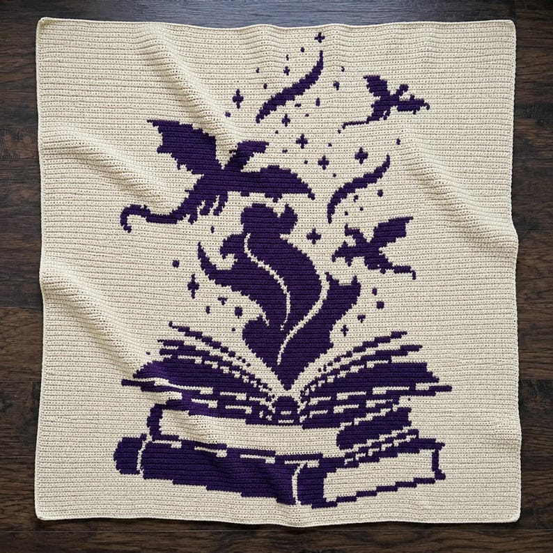 May include: A square crochet blanket in cream with a dark purple graphic design. The pattern features an open book at the bottom with a plume of smoke or magic rising from the pages, surrounded by several small flying dragon silhouettes and stars.