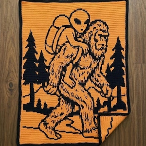May include: Handmade crochet blanket featuring a whimsical design of Bigfoot carrying an alien. The blanket is primarily orange with black accents, including the figures and forest silhouettes. The border is black.