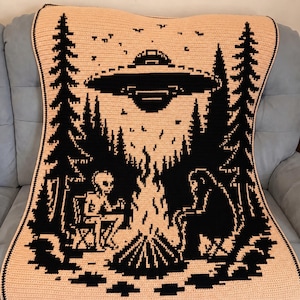 May include: A rectangular crochet blanket featuring a two-tone black and peach design. The graphic depicts an alien and a sasquatch sitting by a campfire in a forest, with a large UFO hovering in the night sky above the trees and small stars.