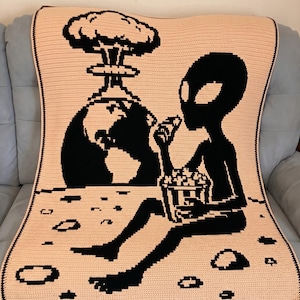 Op de afbeelding: Een perzikgekleurde gehaakte deken met een zwarte alien die popcorn eet en een paddenstoelwolk boven de aarde bekijkt. De deken heeft een zwarte rand en een pixelachtig ontwerp, perfect voor een uniek woondecoratie-item.