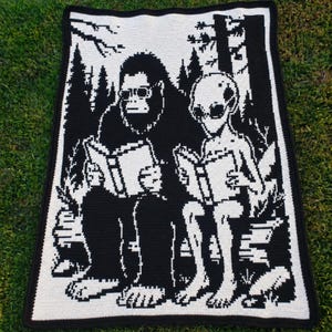 Può includere: Una coperta rettangolare all'uncinetto con un design grafico in bianco e nero che raffigura una grande creatura pelosa e un alieno seduti fianco a fianco in un bosco, entrambi con libri aperti in mano. Bordo nero solido lungo il perimetro.