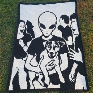 May include: Black and white crochet blanket featuring an alien holding a dog, surrounded by people. The blanket has a pixelated design, with the figures and dog in white against a black background. The blanket is rectangular.