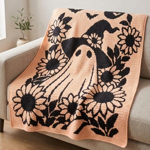 May include: A peach-coloured crochet blanket featuring a large central ghost wearing a witch hat, surrounded by black sunflower motifs and small bats. The textile design uses a contrasting black and peach colour scheme with a repeating floral pattern.