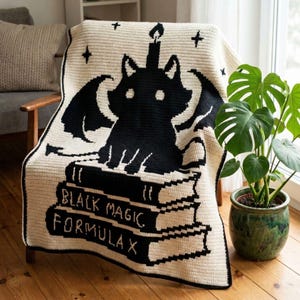 Puede incluir: Una manta de ganchillo color crema con un gato negro con alas de murciélago, una vela en la cabeza y una pila de libros. Los libros dicen "BLACK MAGIC FORMULA X". Las estrellas están esparcidas por la manta.