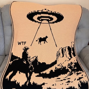 May include: A crocheted blanket with a black and peach design. The blanket shows a UFO abducting a cow, a cowboy on a horse, and the text "WTF". The blanket has a black border.