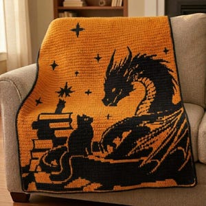 Op de afbeelding: Oranje en zwarte gehaakte deken met een draak, kat, boeken en sterren ontwerp. De draak staat rechts, met de kat en boeken links. De deken is over een grijze fauteuil gedrapeerd.