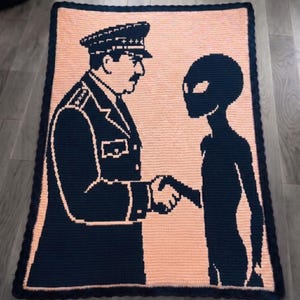 May include: A handmade crochet blanket featuring a navy blue and peach design. The blanket depicts a handshake between a figure in a military uniform and an alien. The border is navy blue.