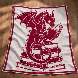 May include: A rectangular crochet blanket featuring a white background with a deep red border and a central graphic design. The design depicts a dragon with wings spread, sitting atop a stack of books while reading an open book in its claws.