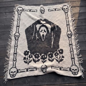 May include: A rectangular cream-colored crochet blanket featuring a central black graphic of a masked figure with hands raised. The border consists of black skulls and bones, with additional skulls and small flowers decorating the design.
