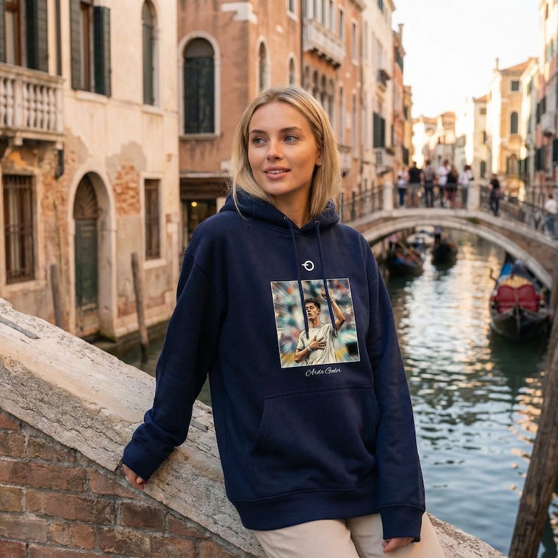 Puede incluir: Sudadera con capucha azul marino con un gr&aacute;fico de una persona. La sudadera tiene una capucha con cord&oacute;n y un bolsillo de canguro. El fondo muestra un puente y un canal.