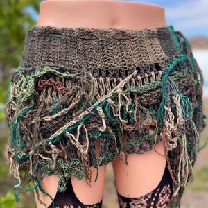 May include: A handmade crochet skirt in shades of green, brown, and beige. The skirt features a textured, open weave design with fringe detailing. The skirt is displayed on a mannequin, showcasing its unique, bohemian style.