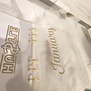 May include: A clear, plastic planner insert with gold lettering that reads "January" and "Life". The planner has gold-colored metal rings. The background is a white marble pattern.