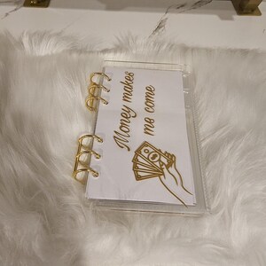May include: A clear, small binder with gold-colored rings and accents. The binder contains a white page with the text "Money makes me come" and a graphic of a hand holding money. The binder is on a white, fluffy surface.