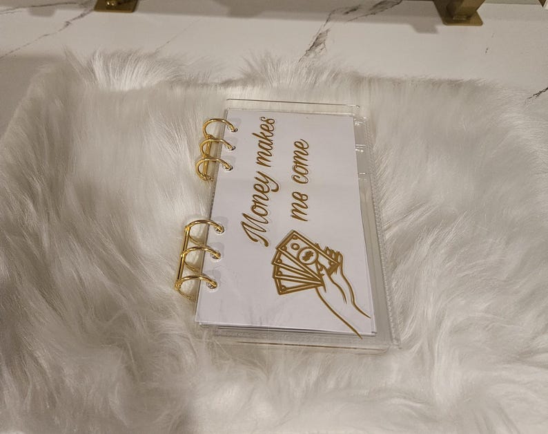 May include: A clear, small binder with gold-colored rings and accents. The binder contains a white page with the text "Money makes me come" and a graphic of a hand holding money. The binder is on a white, fluffy surface.