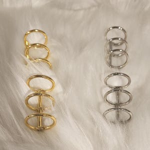 May include: Two sets of metal rings, one gold and one silver, are displayed against a white, fluffy background. Each set consists of four connected rings, creating a unique design. The gold rings have a warm, shiny finish, while the silver rings have a cool, metallic sheen.