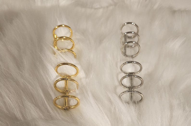 May include: Two sets of metal rings, one gold and one silver, are displayed against a white, fluffy background. Each set consists of four connected rings, creating a unique design. The gold rings have a warm, shiny finish, while the silver rings have a cool, metallic sheen.