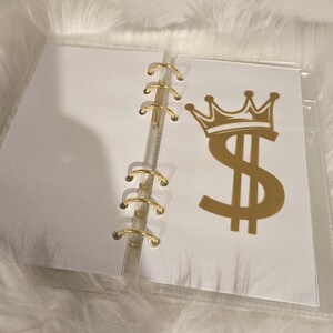 May include: A clear, binder-style organizer with gold-colored rings. One page features a gold dollar sign with a crown. The binder is open, revealing blank white pages.