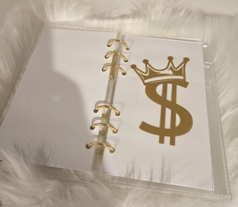 May include: A clear, binder-style organizer with gold-colored rings. One page features a gold dollar sign with a crown. The binder is open, revealing blank white pages.