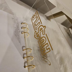 May include: Clear plastic binder with gold-colored metal rings. The binder has a clear plastic cover with the word "Hustle" in gold lettering. The binder is designed for organizing papers and documents.