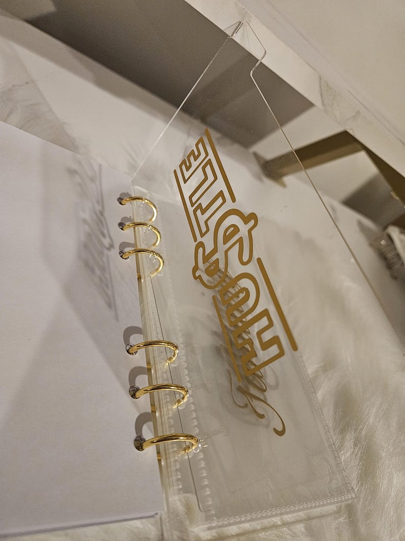 May include: Clear plastic binder with gold-colored metal rings. The binder has a clear plastic cover with the word "Hustle" in gold lettering. The binder is designed for organizing papers and documents.