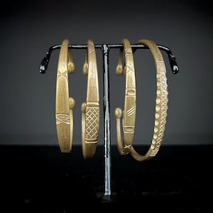 Puede incluir: Cuatro pulseras antiguas de latón con diseños intrincados se exhiben en un soporte de metal negro. Las pulseras presentan varios patrones, incluyendo formas geométricas y superficies texturizadas. Las pulseras son de un color dorado cálido, y el fondo es oscuro.