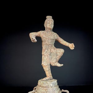 May include: A weathered bronze statue of a dancer in a dynamic pose, balanced on a pedestal supported by elephant figures. The dancer wears detailed traditional attire. The statue has a vintage aesthetic and is set against a black background.